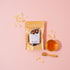 Packaged product with a label on a pink background, surrounded by dried fruit pieces and a small bowl of honey.
